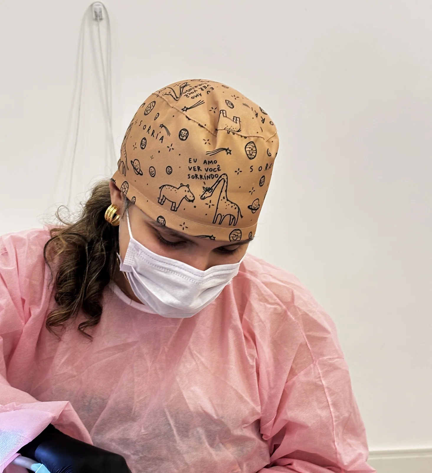 Dra. Gi de máscara e avental rosa durante um atendimento, com a touca caramelo estampada de girafas e planetas onde está escrito à mão “Eu amo ver você sorrindo”